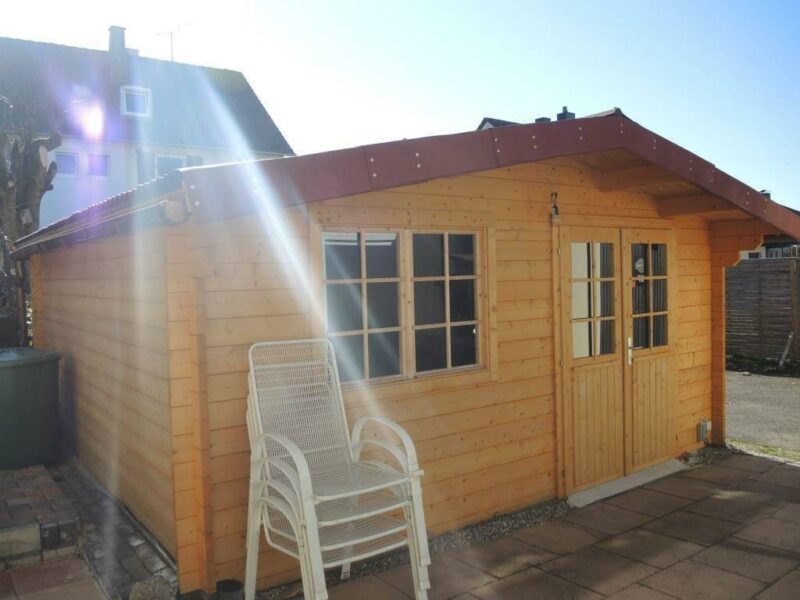Gartenhaus versetzen: Ansicht einer Gartenhütte aus Holz von vorne, davor stapeln sich weiße Gartenstühle.