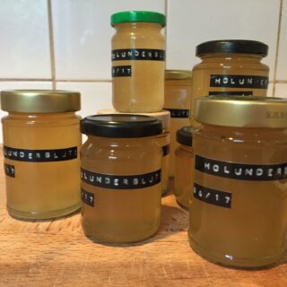 Auf einem Holzbrett stehen mehrere Gläser mit Holunderblütengelee, die mithilfe eines Dymo-Gerätes beschriftet wurden.