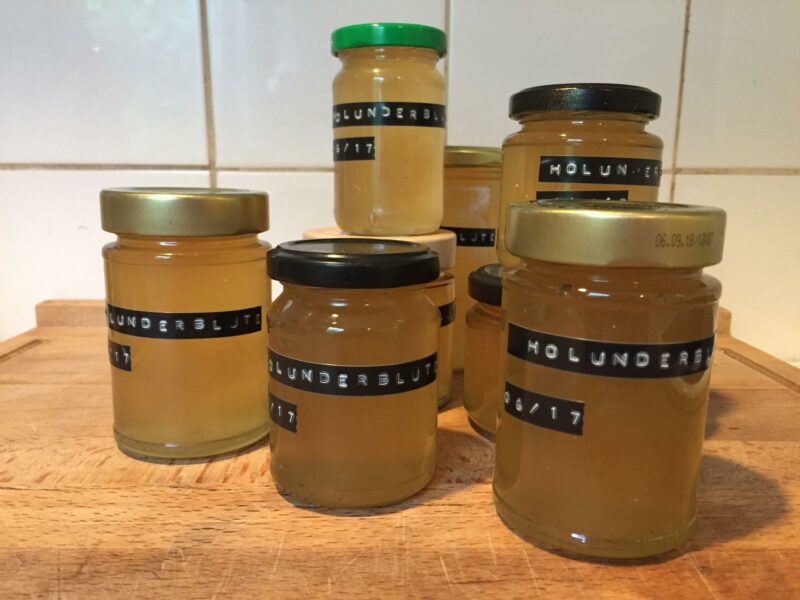 Auf einem Holzbrett stehen mehrere Gläser mit Holunderblütengelee, die mithilfe eines Dymo-Gerätes beschriftet wurden.