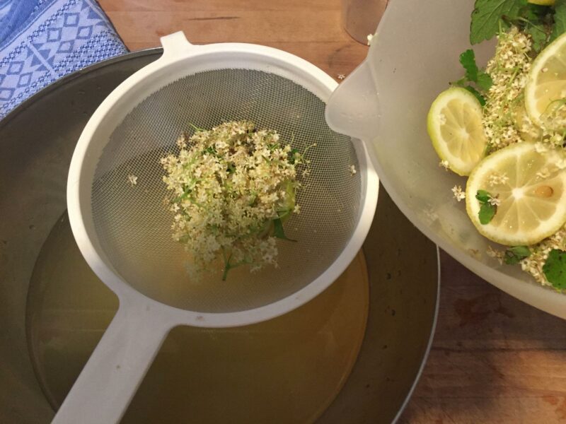 Eine Schüssel mit einem eingehängten Sieb steht auf einem Holzbrett, von rechts wird aus einer weiteren Schüssel Sud von Holunderblüten und Zitronen hineingegossen.