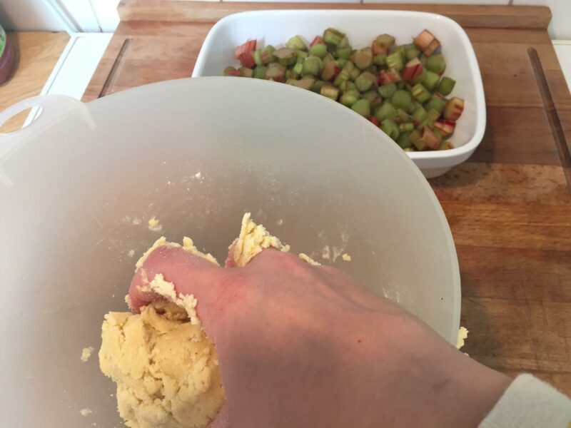 Eine Hand knetet Streuselteig für einen Rhabarbercrumble, im Hintergrund steht eine Auflaufform mit Rhabarber-Stückchen.