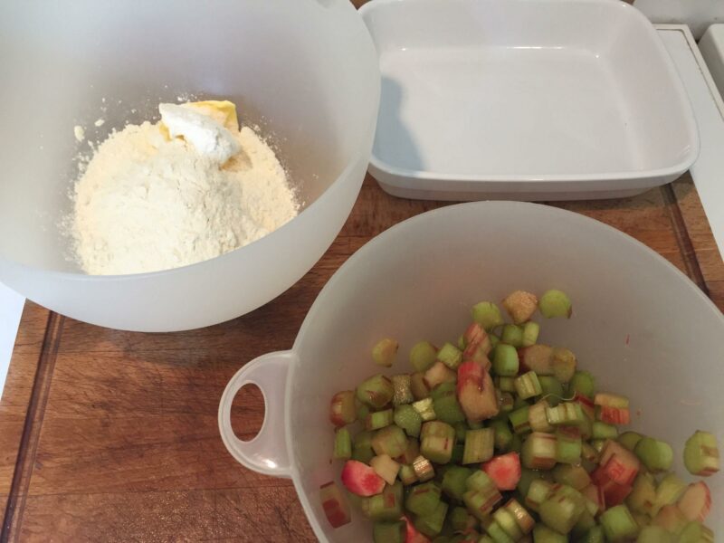 Zutaten für Rhabarbercrumble stehen auf einem Holzbrett in Schüsseln bereit, im Hintergrund steht eine Auflaufform.