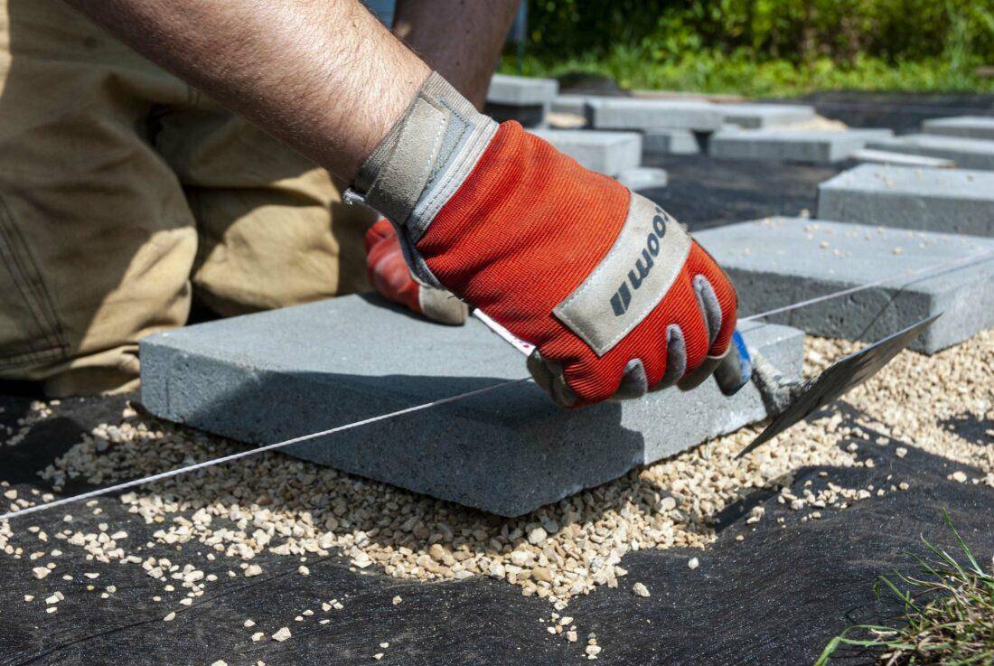 Ein Mann mit roten Gartenhandschuhen verlegt Gehwegplatten als Fundament für eine selbstgebaute Holzterrasse.