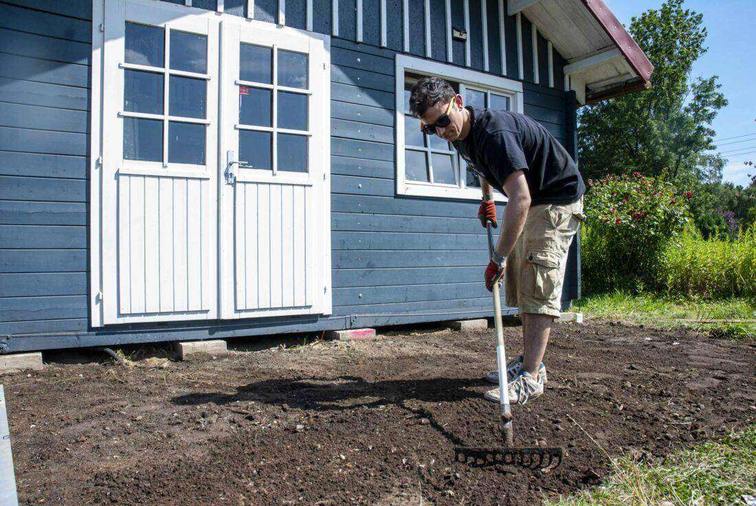 Holzterrasse bauen: Ein Mann mit einer Harke ebnet den Boden vor einer Gartenhütte. 