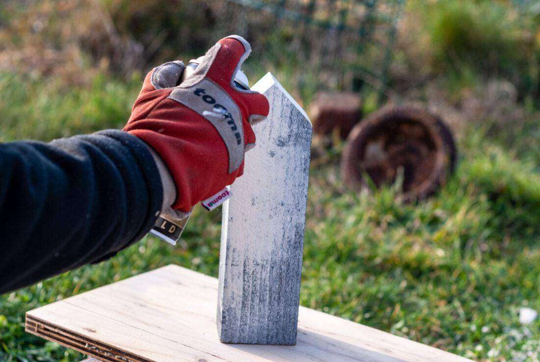 Detailansicht einer Hand eines Mannes, der ein Dekohaus aus Holz mit Lack besprüht.