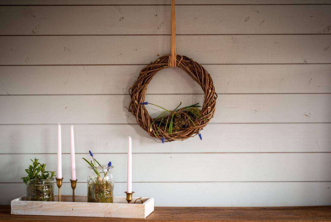 Osterdeko selber machen: Schale mit bepflanzten Weckgläsern und Kerzen vor einer Wand, an dem ein Türkranz hängt.