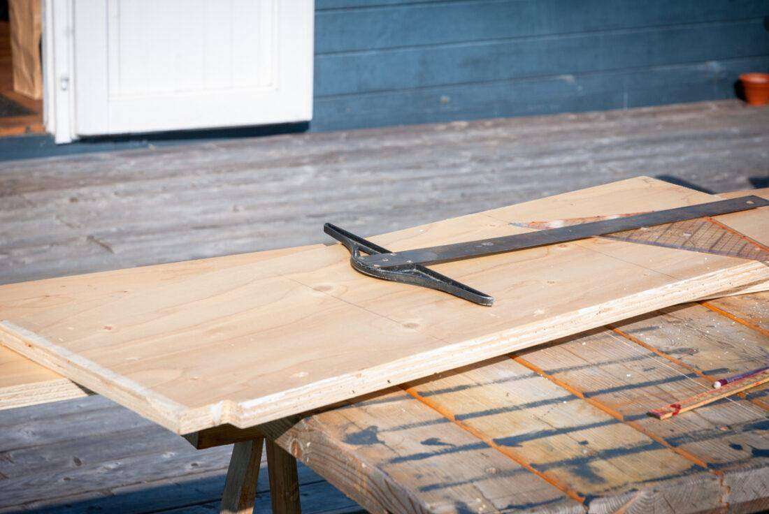 Ein Stück Sperrholz mit Messinstrumenten lieg auf einer Arbeitsplatte vor einer Holzterrasse. 