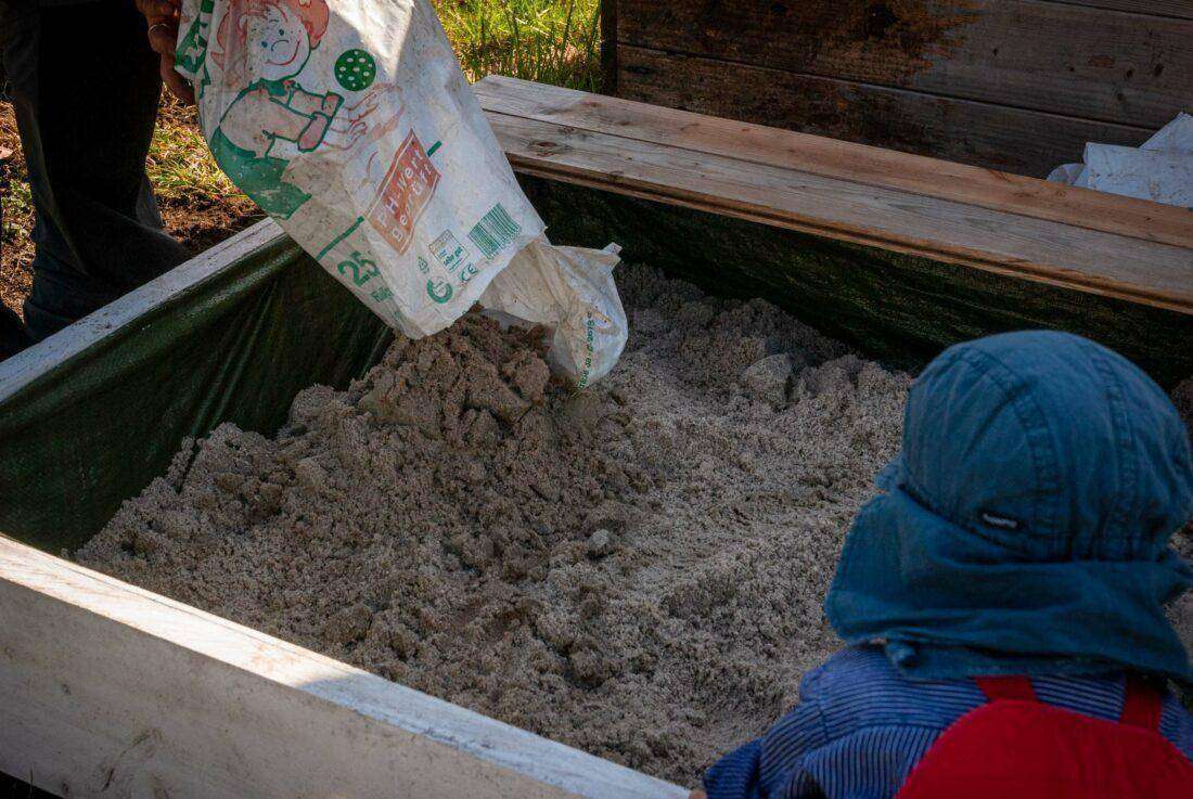 Sandkasten selber bauen: ein Mann füllt Sand in einen Sandkasten, während ein kleines Kind ihm zuguckt. 