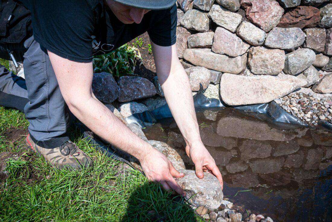 Teich selber bauen: Ein Mann befestigt Teichfolie am Ufer mit einem Stein. 