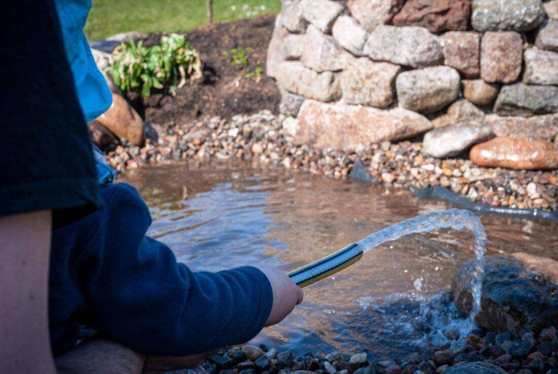 Ein kleines Kind lässt Wasser aus einem Gartenschlauch in einen Teich sprudeln. 