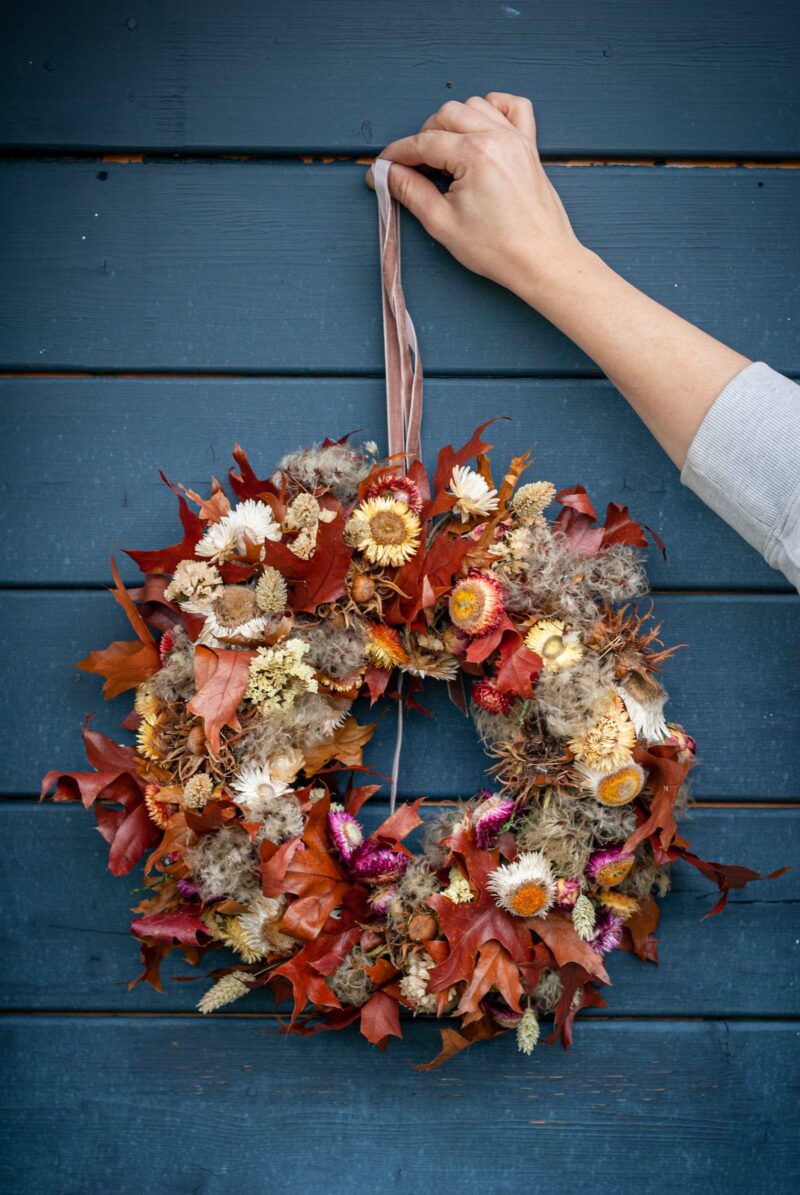 Eine Frau hält einen Herbstkranz vor eine Wand.