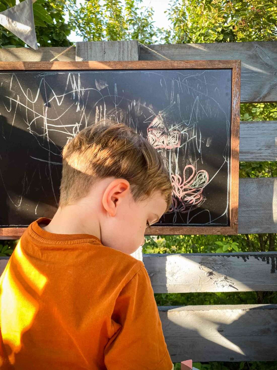 EinJunge sitzt in einem Spielhaus und malt mit Kreide auf eine Tafel.