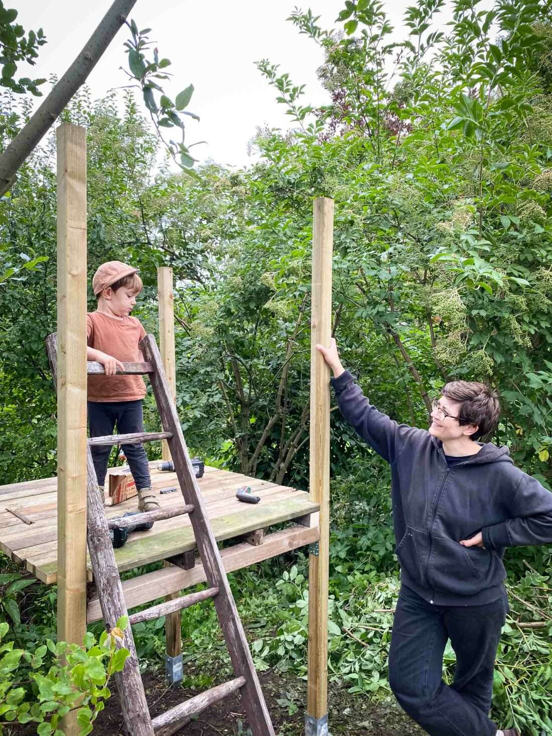 Ein Junge steht auf einem zur Hälfte selbst gebauten Spielhaus. Daneben steht eine Frau und schaut ihn an.