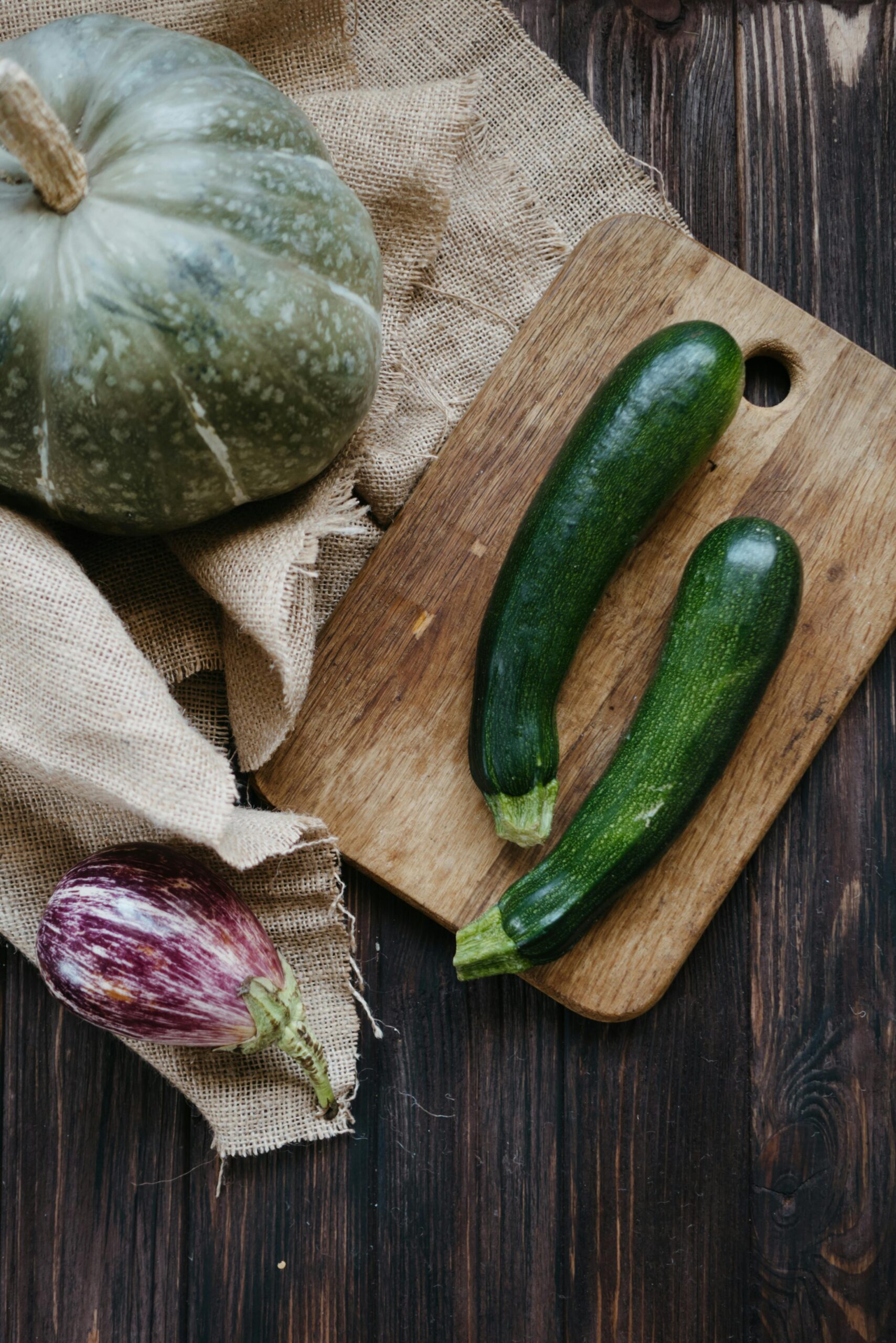 Zucchini-Rezepte. Zwei Zucchini liegen auf einem Holzbrett, daneben liegen ein Kürbis und eine Aubergine.