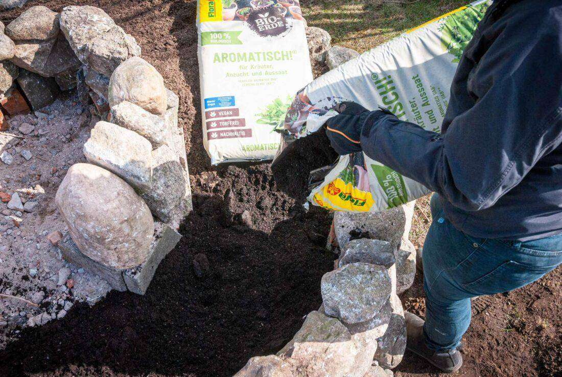 Kräuterspirale mit Erde befüllen: eine Frau verteilt Kräutererde für mediterrane Gewächse. 