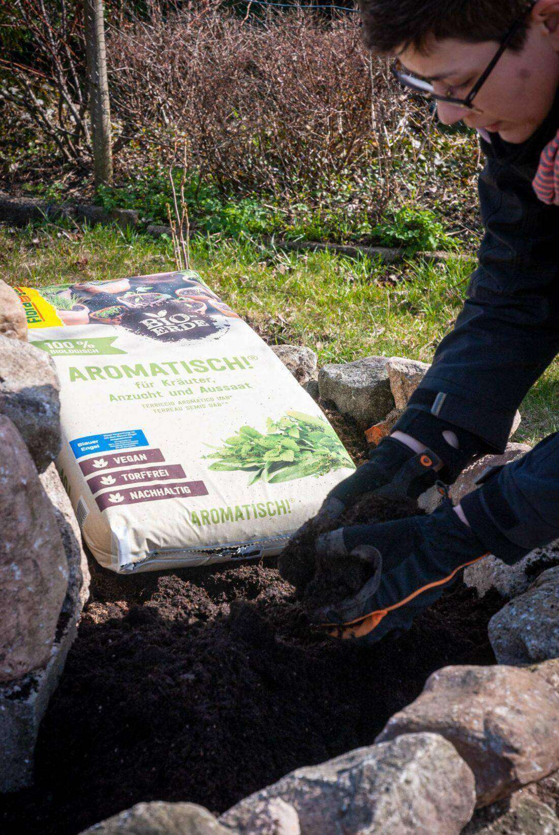 Kräuterspirale mit Erde befüllen: Eine Frau verteilt Erde aus einem Erdsack. 