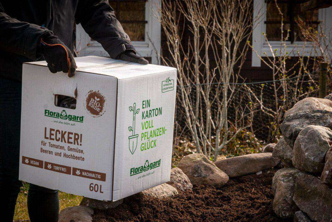 Kräuterspirale mit Erde befüllen: Eine Frau hebt einen Karton Erde auf den Rand einer Kräuterschnecke. 