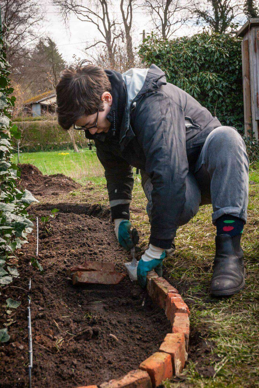 Eine Frau fertig eine Beetumrandung aus Steinen, um einen Sichtschutz um eine Terrasse zu dekorativer in die Gartengestaltung einzubinden. 